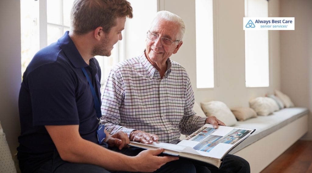 A caregiver and an elderly man are sitting together, looking at a photo album, and sharing a pleasant conversation in a cozy indoor setting.