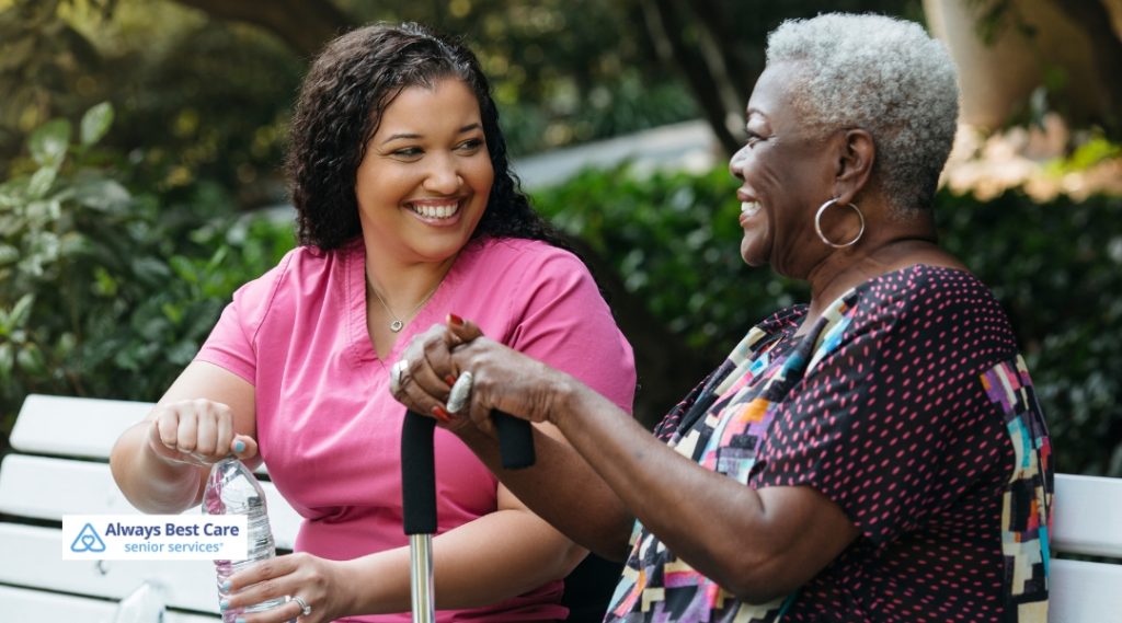A caregiver from Always Best Care is assisting a senior client on a park bench, both smiling and enjoying a pleasant moment in the park.