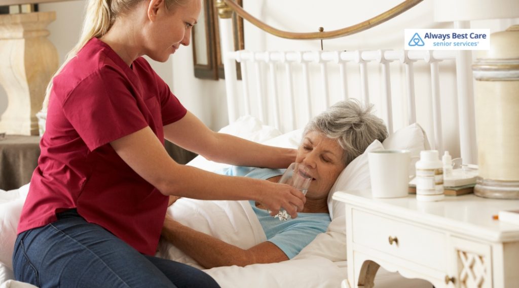 A caregiver in a red scrub shirt is helping an elderly woman in bed, offering her a glass of water with a compassionate, attentive expression.