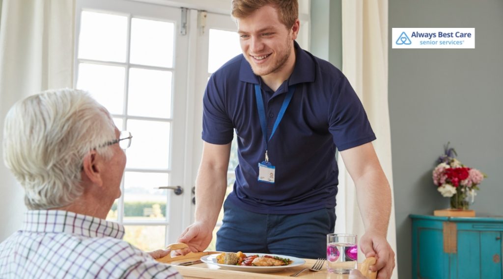 A caregiver in a navy blue shirt is smiling and presenting a meal to an elderly man, sitting at a dining table, offering a sense of warmth and companionship.