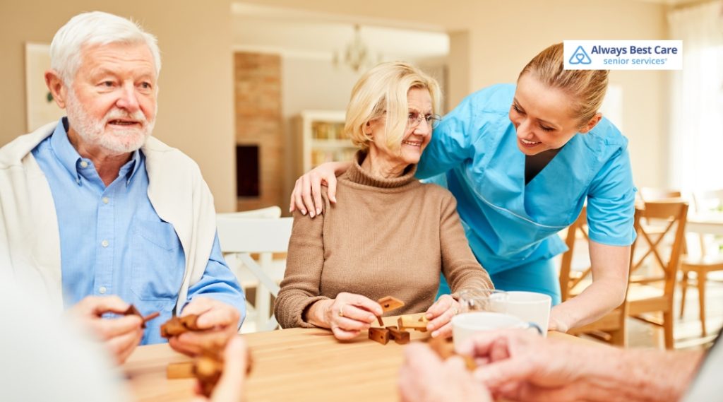 A caregiver in a light blue uniform is helping an elderly woman put on a pink cardigan, with both individuals smiling and showing a caring, nurturing moment.