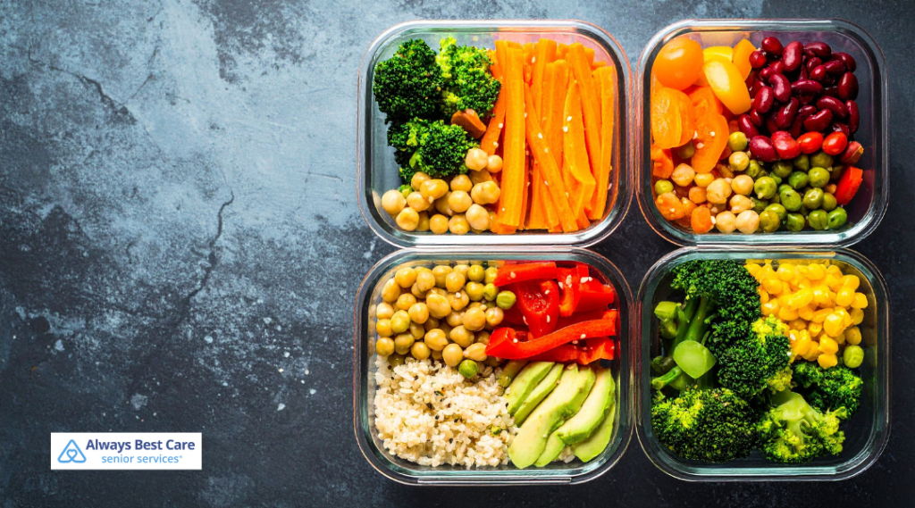 Four clear containers filled with colorful, healthy foods such as vegetables, beans, and grains are arranged on a dark countertop, illustrating meal preparation and nutrition support for seniors. 