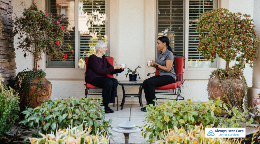 Caregiver and senior woman sitting on a patio, sharing coffee and conversation in a calm outdoor setting.
