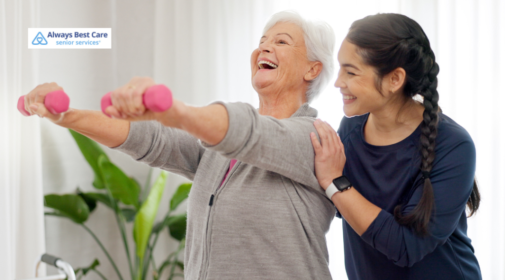Caregiver helping an older woman stand and use a walker beside a hospital bed.