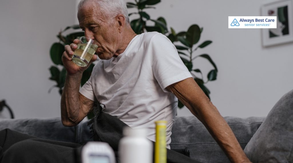 Older man drinking water while seated near medication containers at home.