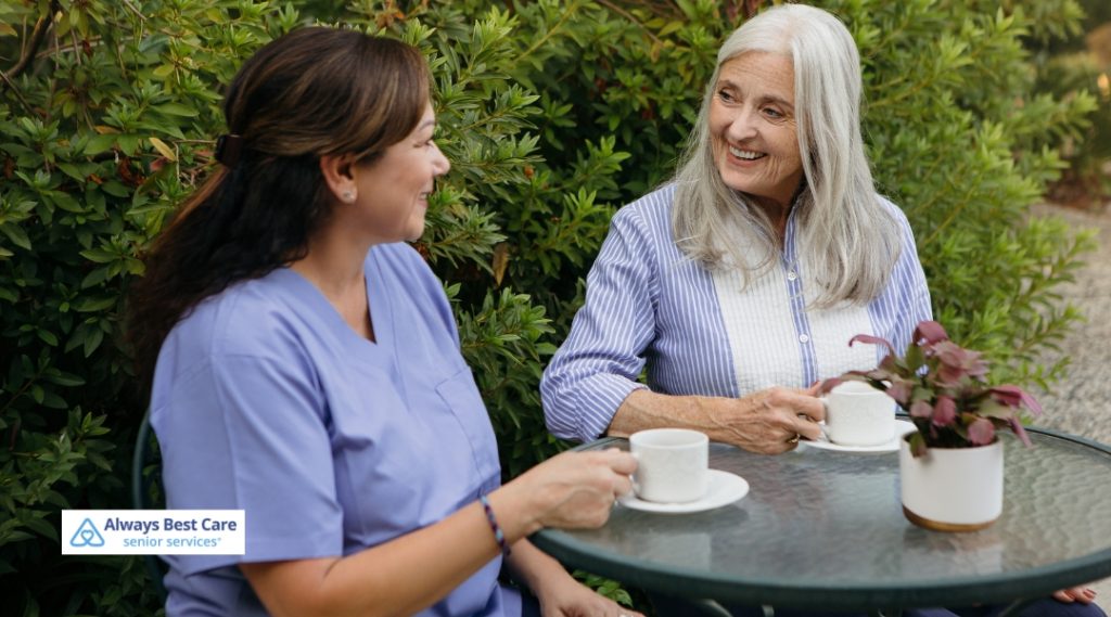 Caregiver and older woman enjoying warm drinks together in a garden.