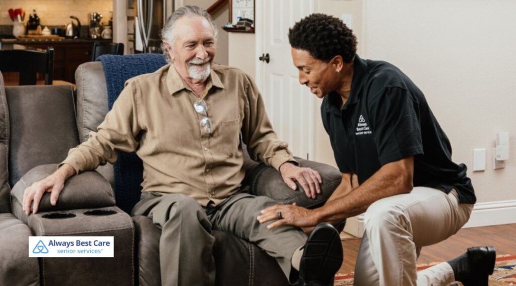 Always Best Care caregiver kneeling beside a smiling senior man in a recliner, checking on his leg during a home care visit.