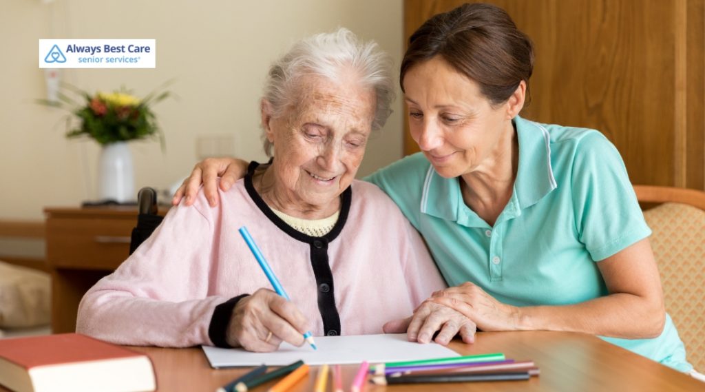 An elderly woman in a wheelchair, wearing a pink sweater, is drawing with colored pencils. A female caregiver in a teal shirt sits beside her, offering support and encouragement with a gentle smile.