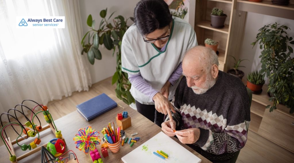 A female caregiver in a white and green uniform assists an elderly man seated at a table. The table is scattered with colorful toys and crafts, suggesting a therapeutic or engaging activity session. The room has bright natural light from a window with sheer curtains.