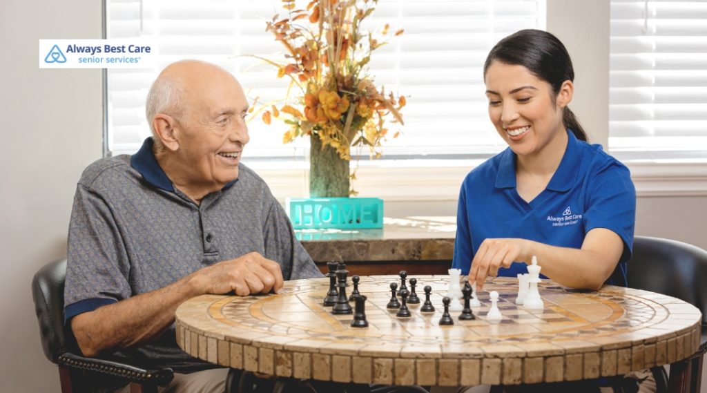 An elderly man and a young female caregiver in a blue polo shirt playing chess together at a round stone table indoors. Both are smiling and enjoying the game, with a window and decorative vase in the background.