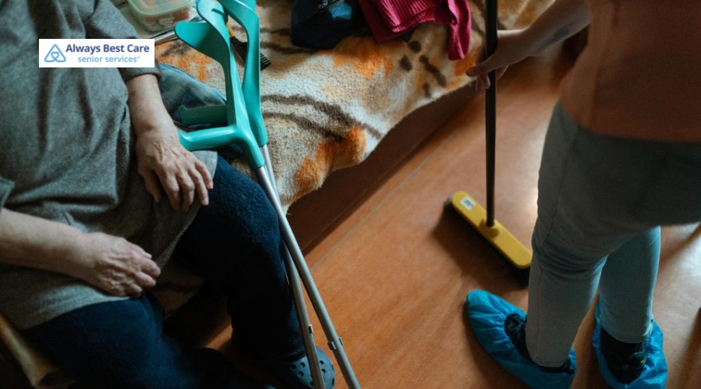 A close-up of an elderly person sitting on a bed with crutches resting beside them, while a caregiver wearing blue shoe covers mops the floor nearby. The scene captures home care assistance with attention to safety and cleanliness.