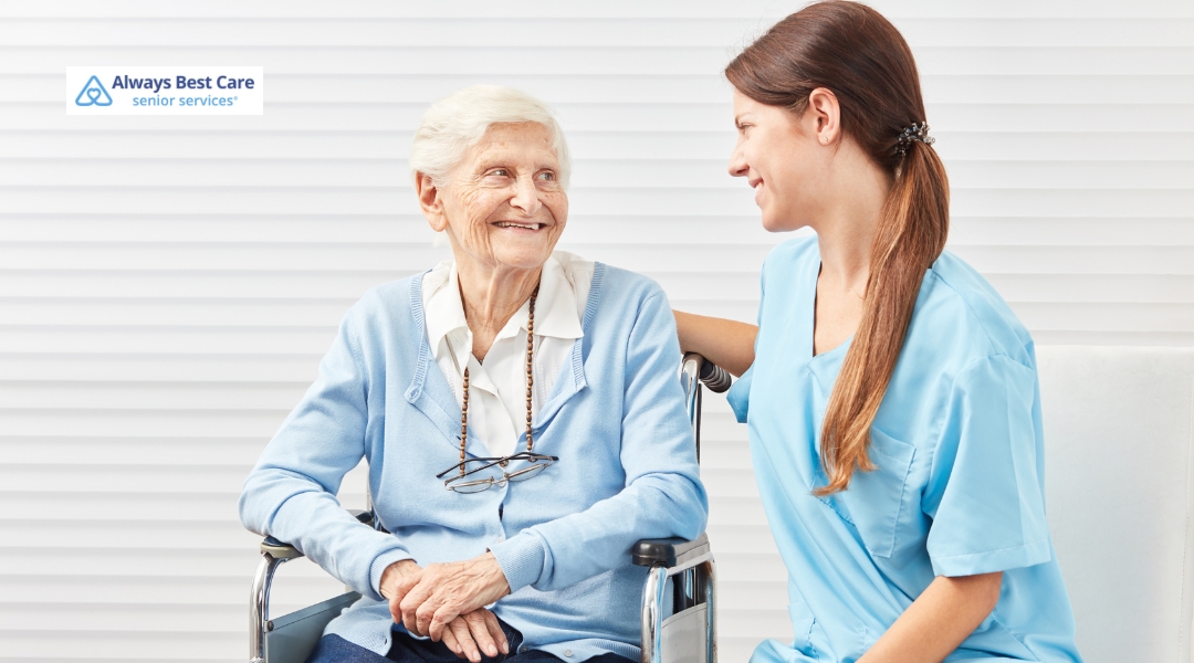 A senior woman is sitting on a couch with a caregiver, enjoying a conversation in a comfortable home setting.