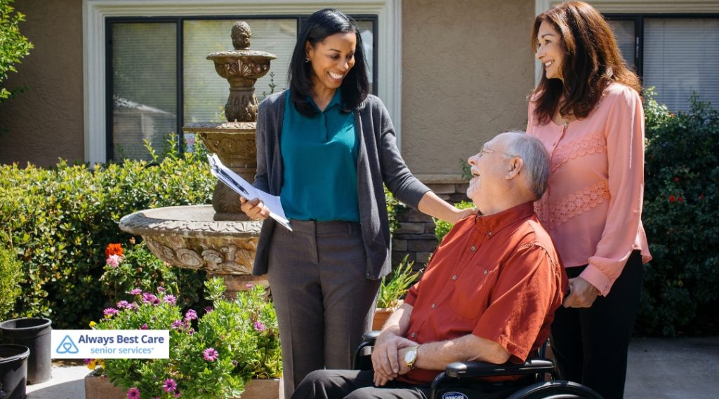 A caregiver is assisting an elderly man in a wheelchair, smiling, while his family members are interacting with him. The Always Best Care logo is in the bottom left corner.