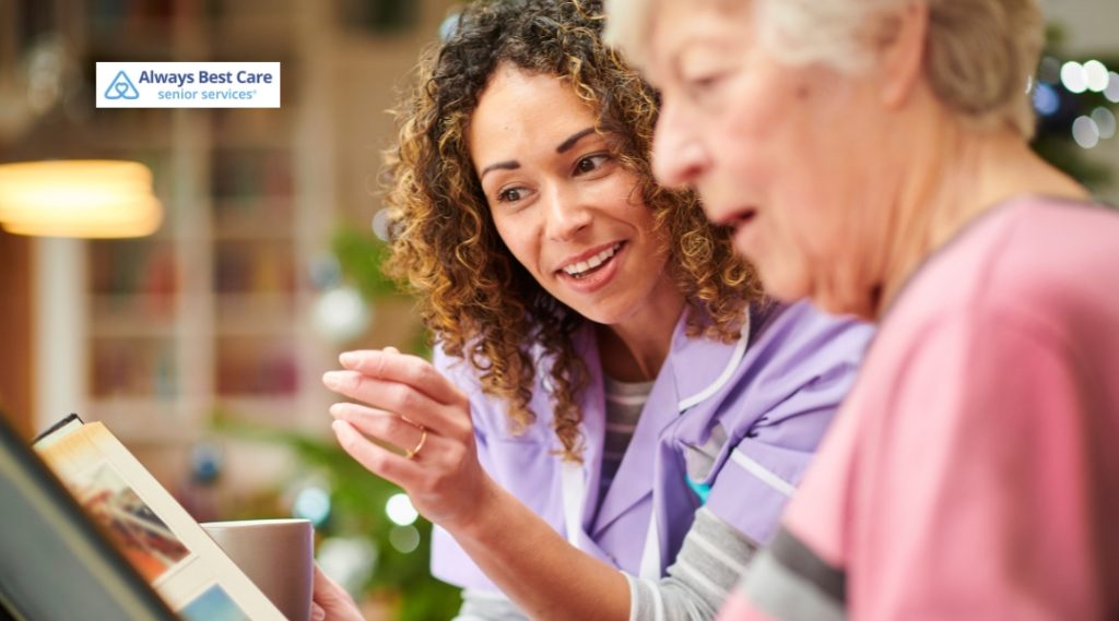 A caregiver in a purple uniform shares a warm moment with an elderly woman while looking at a photo album together. The Always Best Care logo is in the top left corner.