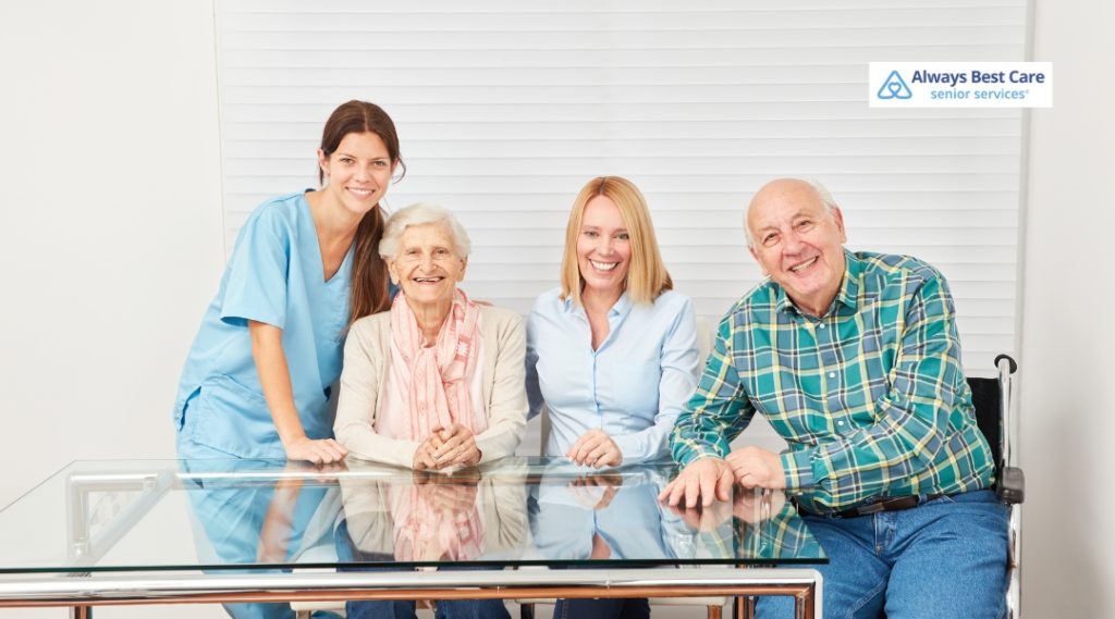 A caregiver in a blue uniform stands beside an elderly woman with gray hair, and they are joined by two family members. The group is smiling and sitting around a glass table, with one family member in a wheelchair.