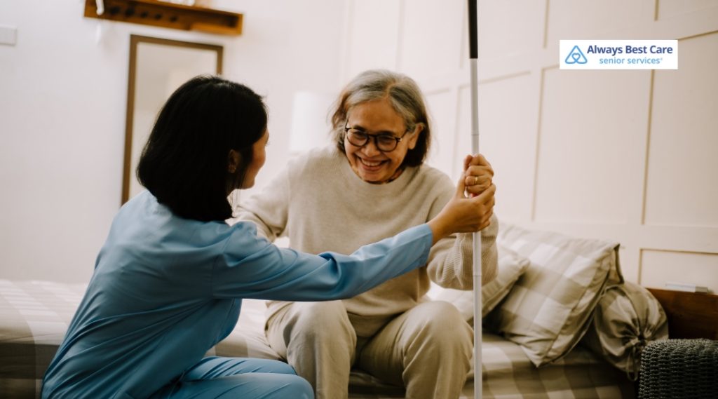 A caregiver in a blue uniform is helping an elderly woman with gray hair and glasses, holding a white cane. The elderly woman is smiling as the caregiver gently assists her.