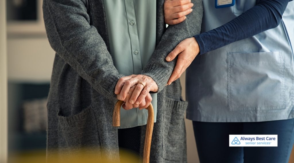 A caregiver in a light blue uniform is assisting an elderly woman with a cane by holding her arm for support. The elderly woman wears a gray sweater and is smiling, standing in a warm, homey setting.