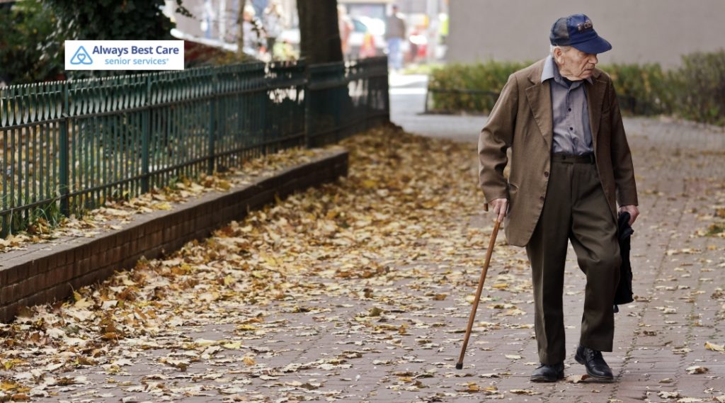 An elderly man walking alone along a leaf-covered sidewalk with a cane. He appears lost or disoriented. The Always Best Care logo is located in the top left corner.