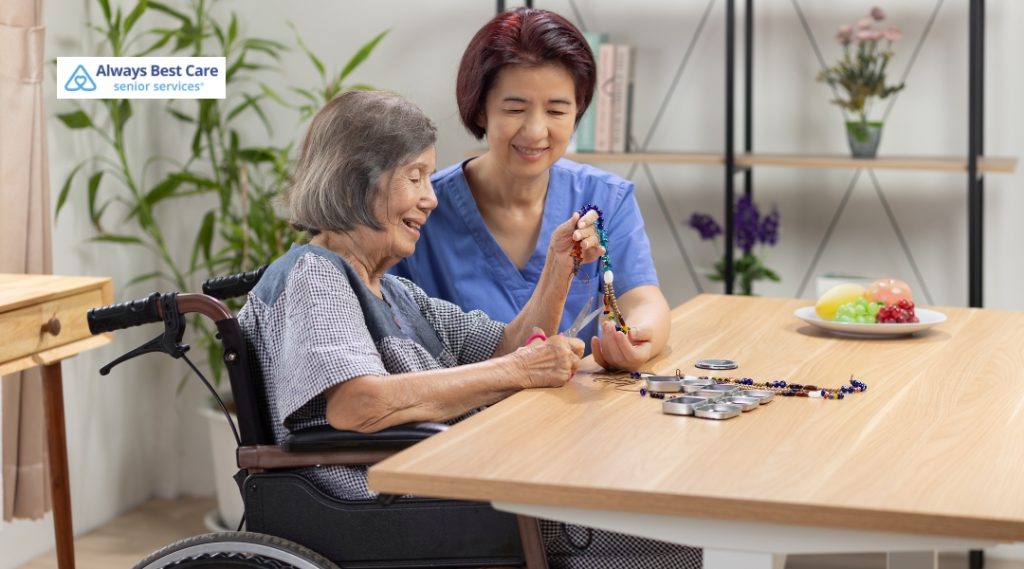 A smiling elderly woman in a wheelchair creates a colorful beaded craft with help from a cheerful caregiver in blue scrubs at a well-lit table.