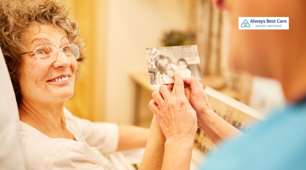 An elderly woman smiles warmly while holding a black-and-white photo shown to her by a caregiver, engaging in memory stimulation or reminiscence therapy.