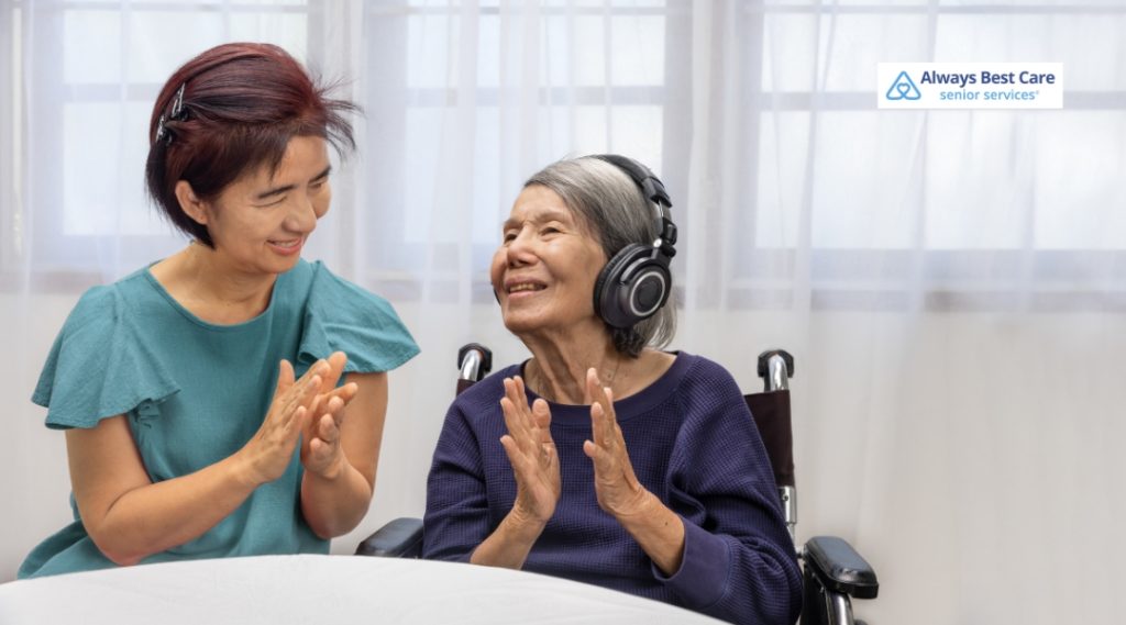 A joyful senior woman wearing headphones claps along with music as a smiling caregiver joins in, promoting cognitive engagement through music therapy.