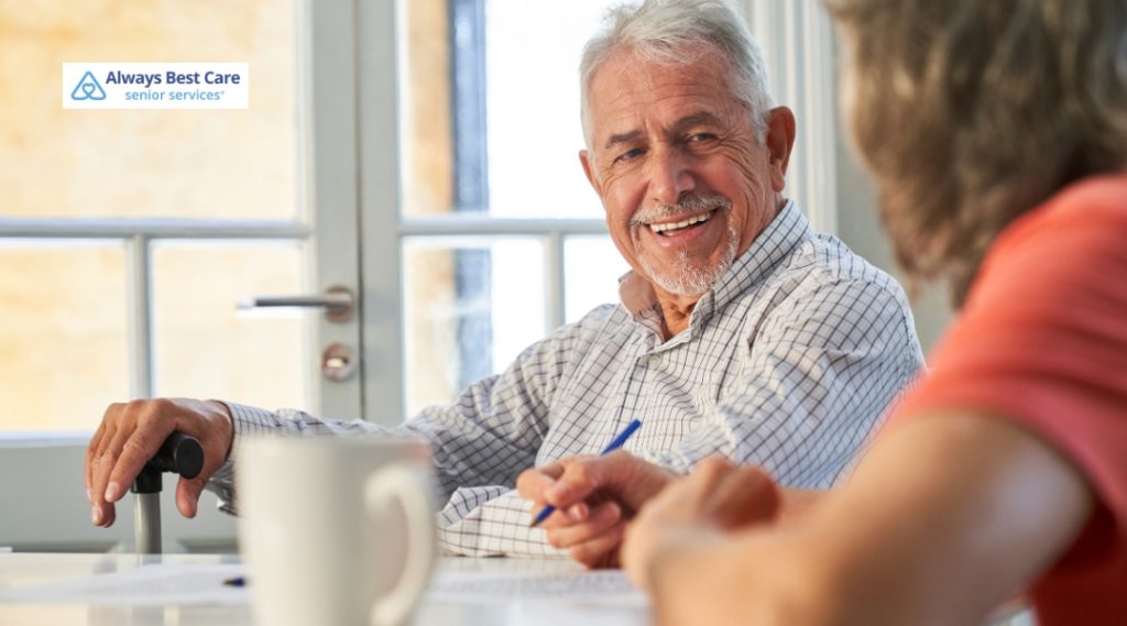 A cheerful elderly man with a cane enjoys a friendly conversation at a table with another person, holding a pen and smiling in a sunlit room.