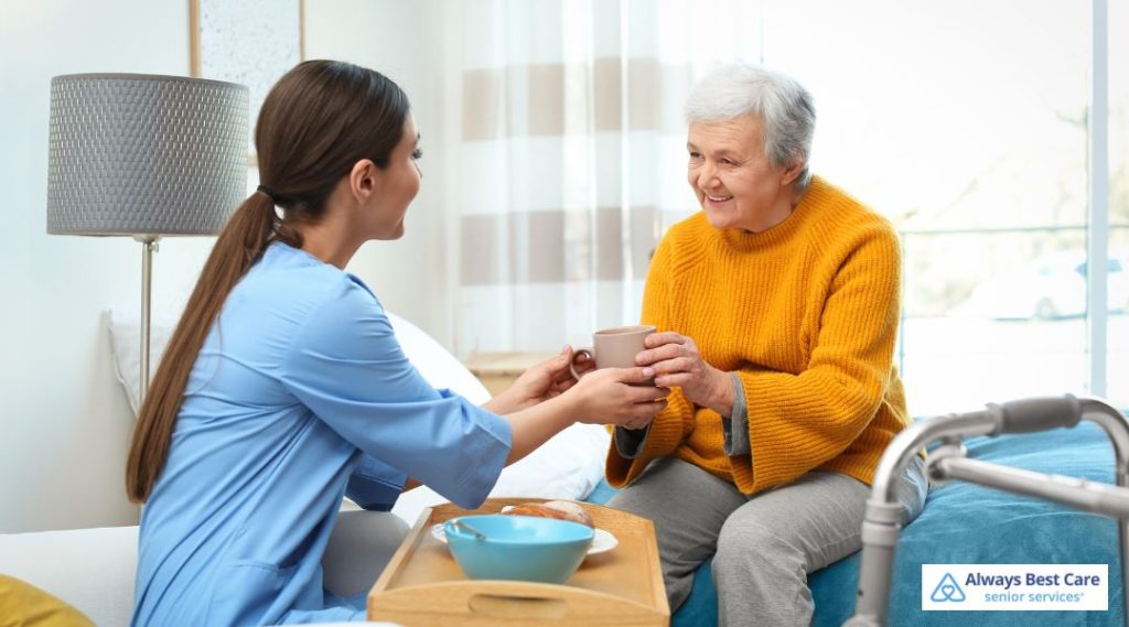 A caregiver in blue scrubs hands a warm cup to a senior woman in a mustard yellow sweater, both smiling warmly while seated near a tray with breakfast items.
