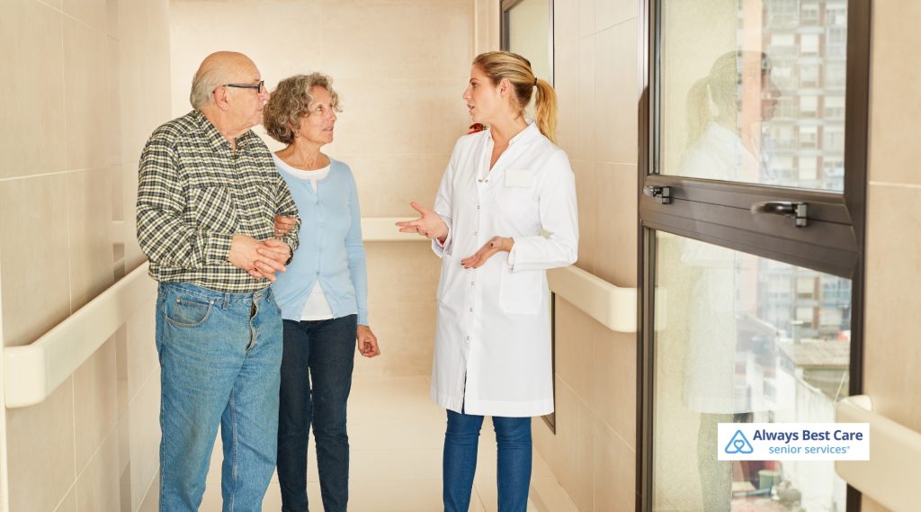 A medical professional in a white coat speaks compassionately with an elderly couple in a modern hallway.