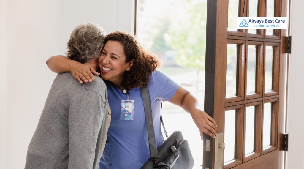 A female caregiver in blue Always Best Care scrubs greets an older man with a warm hug at his open front door.