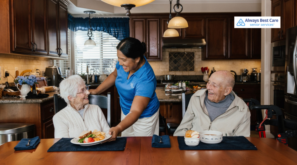 Caregiver serving a healthy meal to two smiling seniors seated together at a kitchen table.