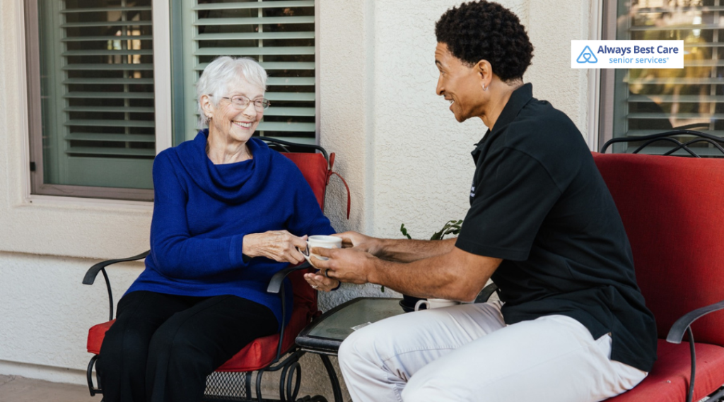Caregiver handing a warm drink to a senior woman while they relax together on an outdoor patio.