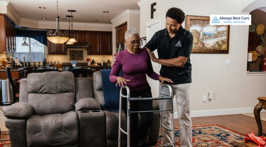 Caregiver supports an older woman using a walker in a living room near a recliner, with a kitchen in the background; ‘Always Best Care senior services’ logo in the corner.