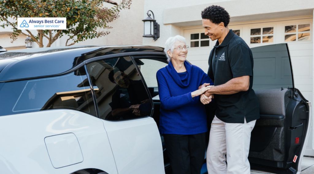 Caregiver helps an older woman step out of a white car in a driveway; both smiling beside the open door; ‘Always Best Care senior services’ logo in the corner.”
