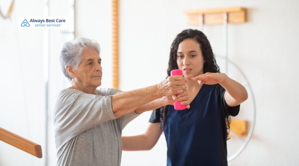 An older woman lifts a small weight while a therapist guides her arm during a rehabilitation session