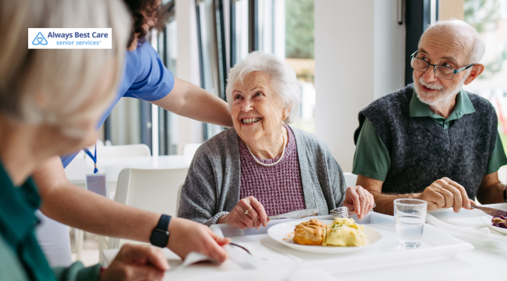 Smiling older adults enjoying a meal together with help from a caregiver.