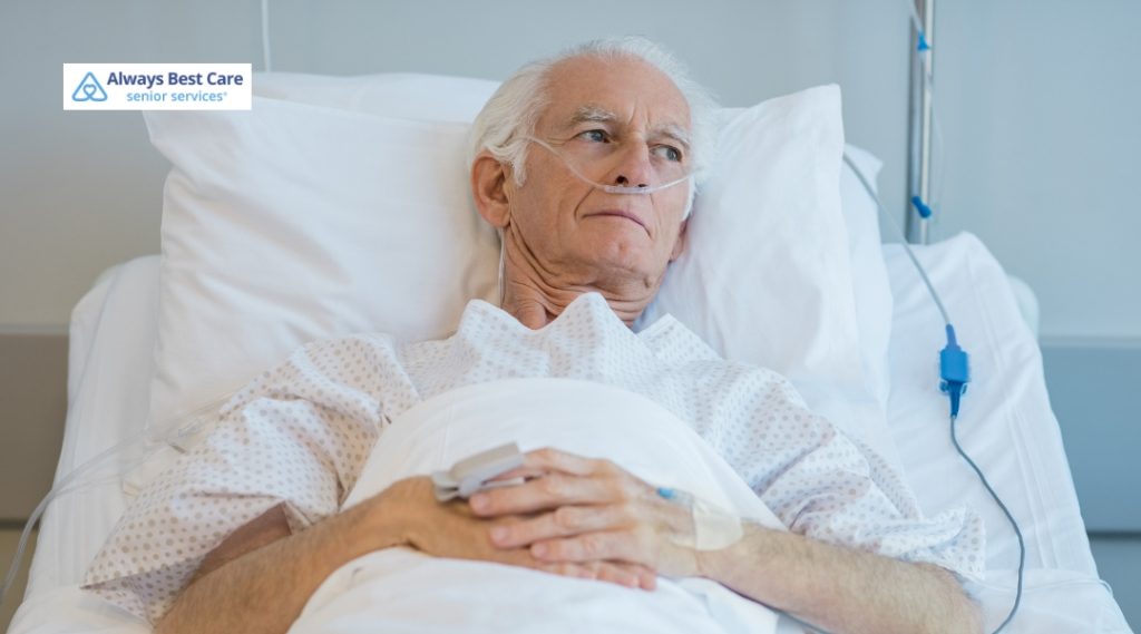 An elderly man with white hair lies in a hospital bed wearing a patient gown and an oxygen nasal cannula. He looks thoughtfully toward the distance while resting with his hands folded on his chest.