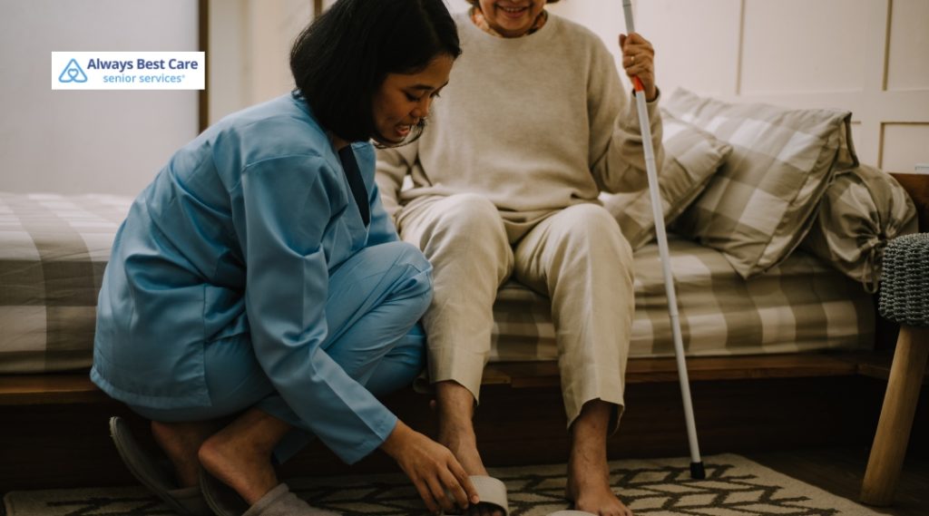 A female caregiver kneels beside an elderly woman seated on a bed indoors. The caregiver is helping the woman put on a slipper, while the woman holds a white cane and smiles. The room has soft natural lighting and a cozy atmosphere.
