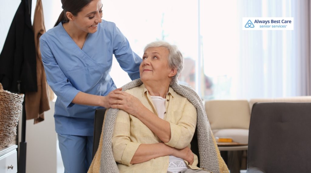 A smiling female caregiver in blue scrubs stands behind an elderly woman seated in a chair, wrapped in a gray blanket indoors. The caregiver places a comforting hand on the elderly woman's shoulder as they share a gentle moment in a warmly lit living space.