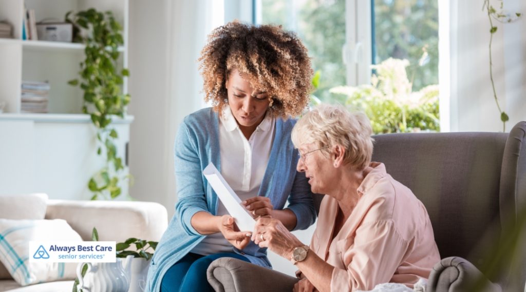 A caregiver with curly hair is showing a photo album to an elderly woman sitting in an armchair. The elderly woman is looking at the album, and both are smiling as they share the moment together.
