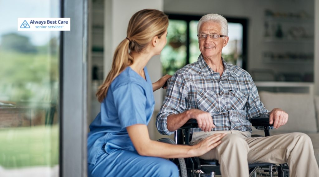 A caregiver in a blue uniform is sitting next to an elderly man in a wheelchair. They are both smiling and enjoying each other's company, while the caregiver offers a comforting presence.