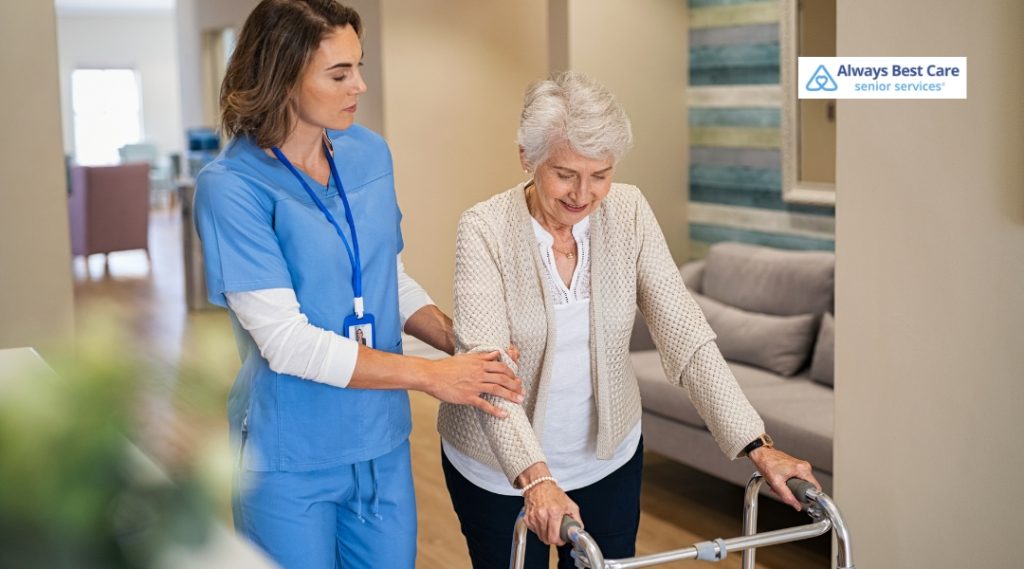 A caregiver in a blue uniform is assisting an elderly woman using a walker, providing support as she takes steps. The elderly woman is smiling as she walks, with the caregiver's hand gently on her back.