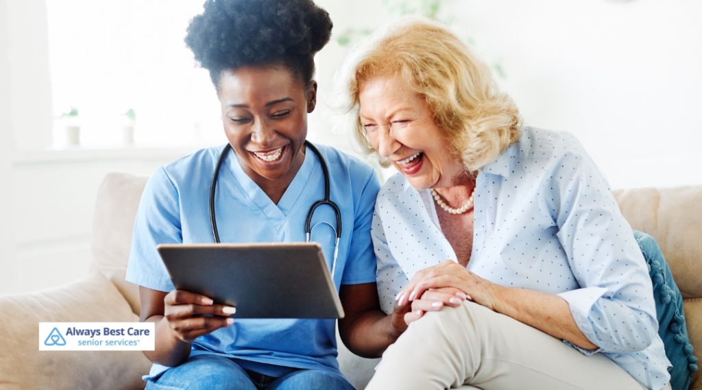 A caregiver in a blue uniform is laughing with an elderly woman while looking at a tablet. The elderly woman is smiling, and both are enjoying a joyful moment together.