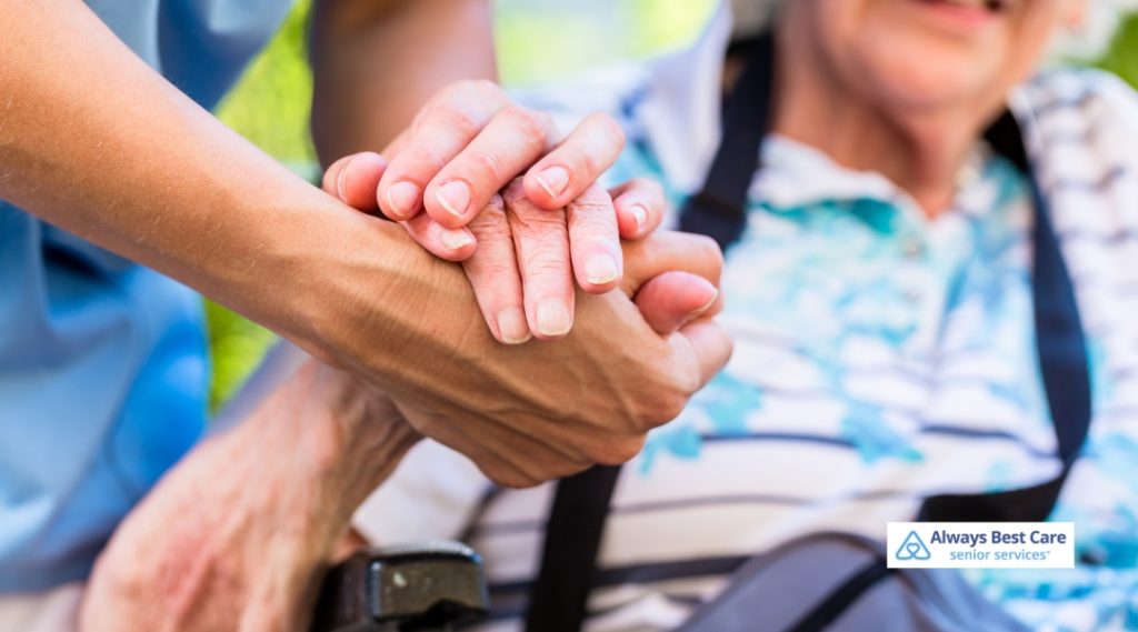 A caregiver in a blue uniform is holding hands with an elderly woman in a wheelchair, offering support and companionship. The caregiver is guiding the elderly woman as they share a tender moment.