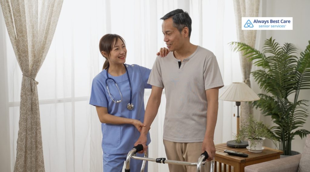 A healthcare worker in blue scrubs supports an elderly man with a walker, smiling and chatting with him in a bright, home-like setting. The caregiver offers a reassuring hand while the man is walking with assistance.