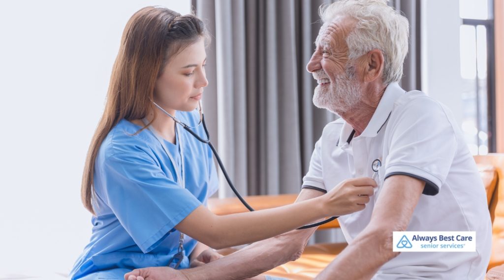 A female healthcare worker checks the blood pressure of an elderly man in a comfortable, light-filled room. The caregiver wears blue scrubs, and the elderly man is smiling as the caregiver uses a stethoscope to listen to his heartbeat.