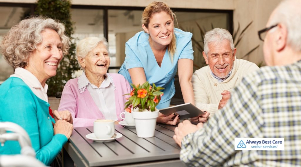 A group of senior adults sits together at a table outside, enjoying coffee and conversation. A caregiver in scrubs smiles as she engages with them, holding a tablet. The scene is cheerful and relaxing, with plants and a sunny patio in the background.