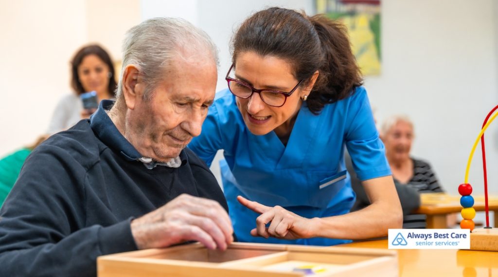 A healthcare worker in blue scrubs helps an elderly man with a game in a therapy setting. The caregiver is pointing at a puzzle as the man carefully engages, with a few other individuals in the background, showcasing a warm, supportive environment.