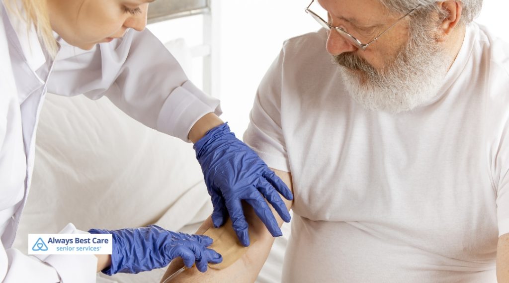 A nurse wearing blue gloves is carefully applying a bandage to the forearm of a senior man dressed in a white t-shirt, providing wound care in a home setting. 
