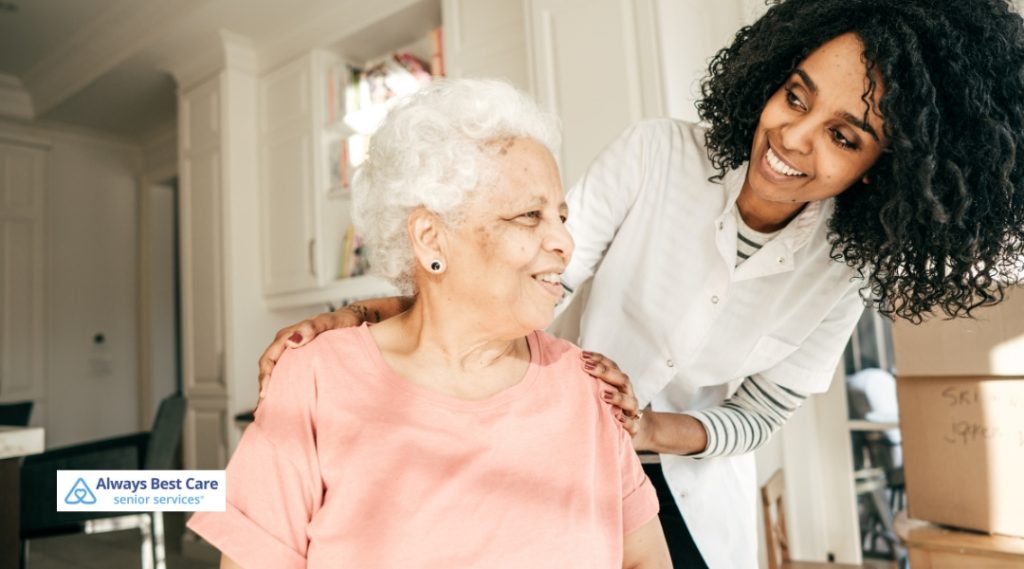 A caregiver stands behind a senior woman dressed in a peach shirt, offering supportive companionship in a bright and comfortable kitchen environment