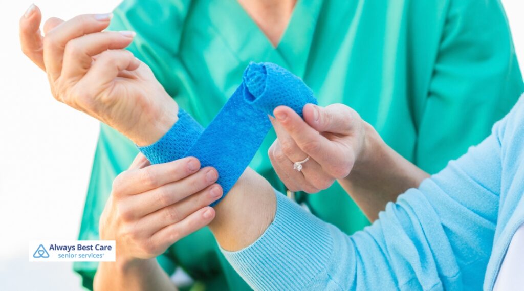 Close-up of a caregiver in green scrubs wrapping a blue cohesive bandage around a patient's wrist and hand for wound protection.